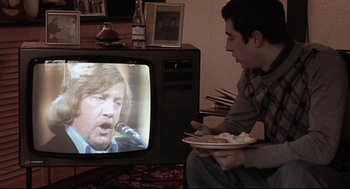 Movie still from “24 Hour Party People” (2002), directed by Tom Bruggen – A man sitting in front of a tv holding a plate of food; Medium shot, High angle