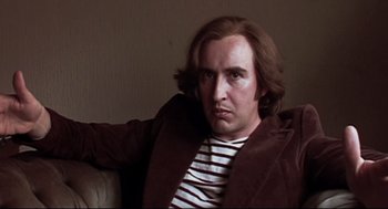 Movie still from “24 Hour Party People” (2002), directed by Tom Bruggen – A man sitting on a couch in a room; Close Up shot, Low angle