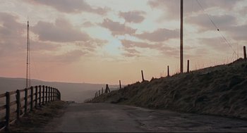 Movie still from “24 Hour Party People” (2002), directed by Tom Bruggen – A car driving down a road next to a fence; Extreme Wide shot, High angle