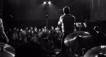 Movie still from “24 Hour Party People” (2002), directed by Tom Bruggen – A man standing on a stage in front of an audience; Wide shot, High angle