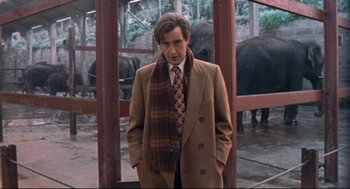 Movie still from “24 Hour Party People” (2002), directed by Tom Bruggen – A man in a suit and tie standing in front of elephants; Medium shot, Low angle