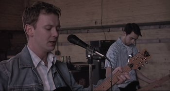 Movie still from “24 Hour Party People” (2002), directed by Tom Bruggen – A man is playing a guitar while another man is singing; Close Up shot, Low angle