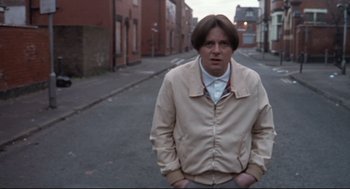 Movie still from “24 Hour Party People” (2002), directed by Tom Bruggen – A man standing on the side of the road in front of some buildings; Medium shot, Low angle
