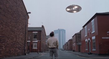 Movie still from “24 Hour Party People” (2002), directed by Tom Bruggen – A man standing on the side of the street looking at a flying saucer; Extreme Wide shot, Low angle