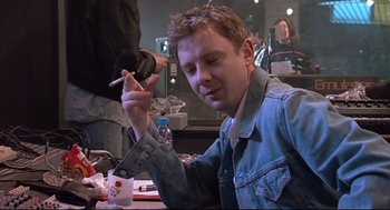 Movie still from “24 Hour Party People” (2002), directed by Tom Bruggen – A man sitting at a table holding a cigarette in his hand; Medium shot, High angle
