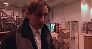 Movie still from “24 Hour Party People” (2002), directed by Tom Bruggen – A man wearing a suit and tie standing in front of boxes in a room; Close Up shot, Low angle