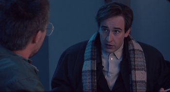 Movie still from “24 Hour Party People” (2002), directed by Tom Bruggen – A man wearing a jacket and a scarf is standing next to another man in a room; Medium shot, Over the shoulder angle