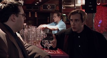 Movie still from “24 Hour Party People” (2002), directed by Tom Bruggen – A man holding out his hand over a bunch of empty glasses; Medium shot, Over the shoulder angle