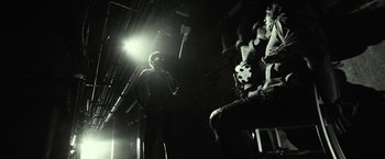 Movie still from “31” (2016), directed by Rob Zombie – A man standing next to another man in a dark room; Wide shot, Over the shoulder angle