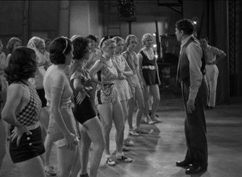 Movie still from “42nd Street” (1933), directed by Lloyd Bacon – A group of women standing next to each other in front of a man; Wide shot, High angle