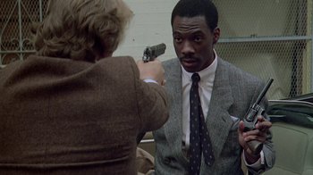 Movie still from “48 Hrs.” (1982), directed by Walter Hill – A man in a suit holding a gun pointed at a woman; Medium shot, Over the shoulder angle