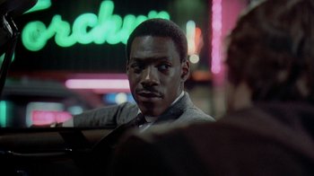 A man in a suit and tie sitting in front of a neon sign