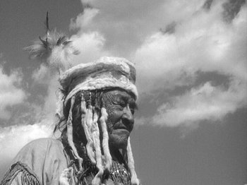 Movie still from “49th Parallel” (1941), directed by Michael Powell – An old man with a feather headdress is standing in front of a cloudy sky; Close Up shot, Low angle