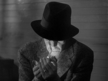 Movie still from “49th Parallel” (1941), directed by Michael Powell – A man in a suit and a hat is smoking a cigarette; Close Up shot, Over the shoulder angle