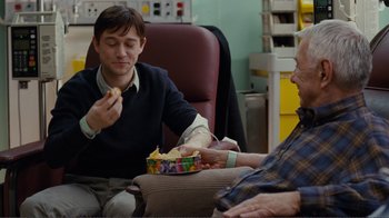 Movie still from “50/50” (2011), directed by Jonathan Levine – A man sitting in a chair eating food from a box; Medium shot, Over the shoulder angle