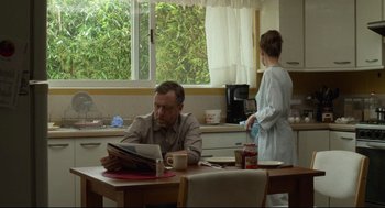 Movie still from “600 Miles” (2015), directed by Gabriel Ripstein – A man sitting at a table reading a newspaper while a woman stands in the background; Medium shot, Over the shoulder angle