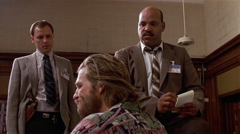 Movie still from “8 Million Ways to Die” (1986), directed by Hal Ashby – A man in a suit and tie is combing another man's hair; Medium shot, Over the shoulder angle