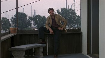 Movie still from “8 Million Ways to Die” (1986), directed by Hal Ashby – A man sitting on top of a bench on a porch; Wide shot, Low angle