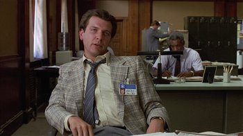 Movie still from “8 Million Ways to Die” (1986), directed by Hal Ashby – A man sitting in front of a desk in an office setting; Medium shot, Low angle