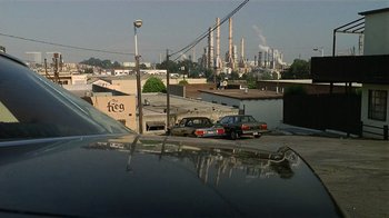 Movie still from “8 Million Ways to Die” (1986), directed by Hal Ashby – Cars are parked on the side of the road near a factory; Extreme Wide shot, Low angle