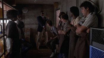 Movie still from “8 Million Ways to Die” (1986), directed by Hal Ashby – A group of people sitting in a room with a man holding a gun in his hand; Wide shot, Over the shoulder angle