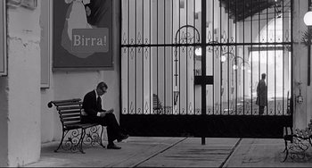 Movie still from “8½” (1963), directed by Federico Fellini – A man sitting on top of a wooden bench outside; Wide shot, Low angle