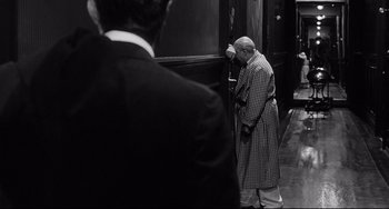 Movie still from “8½” (1963), directed by Federico Fellini – A man in a trench coat and a suit is standing in front of a door; Medium shot, Over the shoulder angle