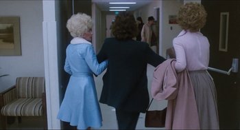 Movie still from “9 to 5” (1980), directed by Colin Higgins – A woman in a blue dress is holding her hand on the back of another woman in a black suit; Wide shot, Over the shoulder angle