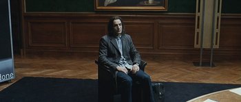 Movie still from “99 Francs” (2007), directed by Jan Kounen – A man in a suit sitting in a chair in front of a painting; Wide shot, High angle