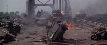Movie still from “A Bridge Too Far” (1977), directed by Richard Attenborough – A car that has been set on fire in the street; Wide shot, High angle