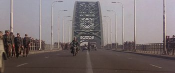 Movie still from “A Bridge Too Far” (1977), directed by Richard Attenborough – A group of people riding motorcycles across a bridge; Extreme Wide shot, High angle