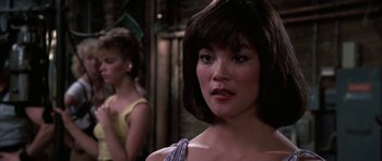 Movie still from “A Chorus Line” (1985), directed by Richard Attenborough – A woman with a short hair cut and bangs looking at the camera with another woman in the background; Close Up shot, Over the shoulder angle