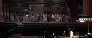 Movie still from “A Chorus Line” (1985), directed by Richard Attenborough – A group of people standing on a stage in front of a brick building; Wide shot, High angle