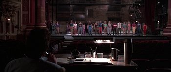 Movie still from “A Chorus Line” (1985), directed by Richard Attenborough – A group of people standing on a stage in front of an audience; Wide shot, Over the shoulder angle
