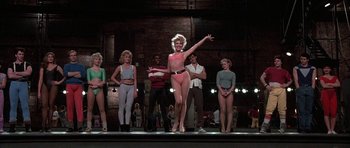 Movie still from “A Chorus Line” (1985), directed by Richard Attenborough – A group of people standing on a stage with a woman in a pink bathing suit; Wide shot, Low angle