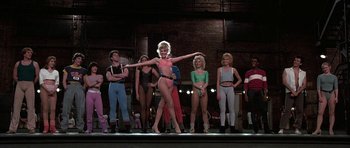 Movie still from “A Chorus Line” (1985), directed by Richard Attenborough – A group of people standing on a stage with a woman in a bikini; Wide shot, Low angle