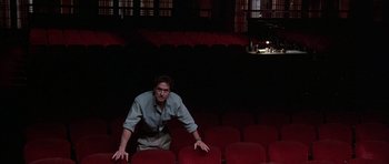 Movie still from “A Chorus Line” (1985), directed by Richard Attenborough – A man sitting in a red chair in front of a piano; Wide shot, Low angle