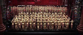 Movie still from “A Chorus Line” (1985), directed by Richard Attenborough – A large group of gold statues in front of a red background; Extreme Wide shot, High angle