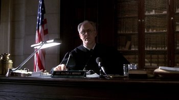 Movie still from “A Civil Action” (1998), directed by Steven Zaillian – A man sitting at a table in front of microphones and an american flag; Medium shot, Low angle