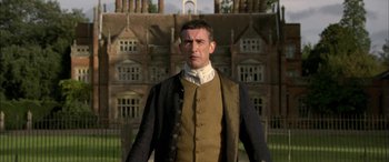 Movie still from “Tristram Shandy” (2005), directed by Michael Winterbottom – A man in a brown and black jacket standing in front of a building; Medium shot, Low angle