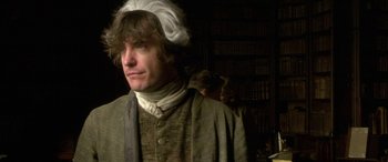 Movie still from “Tristram Shandy” (2005), directed by Michael Winterbottom – A man wearing a hat standing next to a book shelf; Close Up shot, Over the shoulder angle