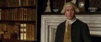 Movie still from “Tristram Shandy” (2005), directed by Michael Winterbottom – A man wearing a wig and a wig stand in front of a fireplace; Medium shot, Over the shoulder angle