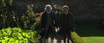 Movie still from “Tristram Shandy” (2005), directed by Michael Winterbottom – A couple of men walking down a path next to flowers; Medium shot, High angle