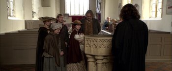 Movie still from “Tristram Shandy” (2005), directed by Michael Winterbottom – A group of people standing in a room next to a stone pillar; Medium shot, Over the shoulder angle