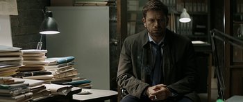 Movie still from “Department Q: A Conspiracy of Faith” (2016), directed by Hans Petter Moland – A man sitting in front of a stack of books; Medium shot, Over the shoulder angle