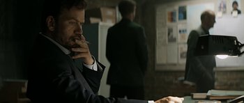 Movie still from “Department Q: A Conspiracy of Faith” (2016), directed by Hans Petter Moland – A man smoking a cigarette in a room with other men; Medium shot, Over the shoulder angle