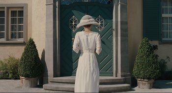 Movie still from “A Dangerous Method” (2011), directed by David Cronenberg – A woman in a white dress and a white hat standing in front of a green door; Wide shot, Over the shoulder angle