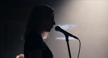 Movie still from “A Date for Mad Mary” (2016), directed by Darren Thornton – A woman standing in front of a microphone in a dark room; Close Up shot, Low angle