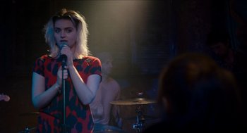 Movie still from “A Date for Mad Mary” (2016), directed by Darren Thornton – A woman holding a microphone while standing in front of a drum set; Medium shot, Over the shoulder angle