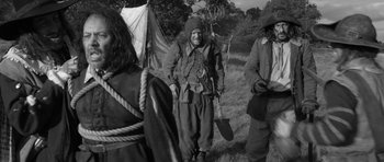 Movie still from “A Field in England” (2013), directed by Ben Wheatley – An old man in a hat and a long beard standing in a field with other people; Wide shot, Over the shoulder angle